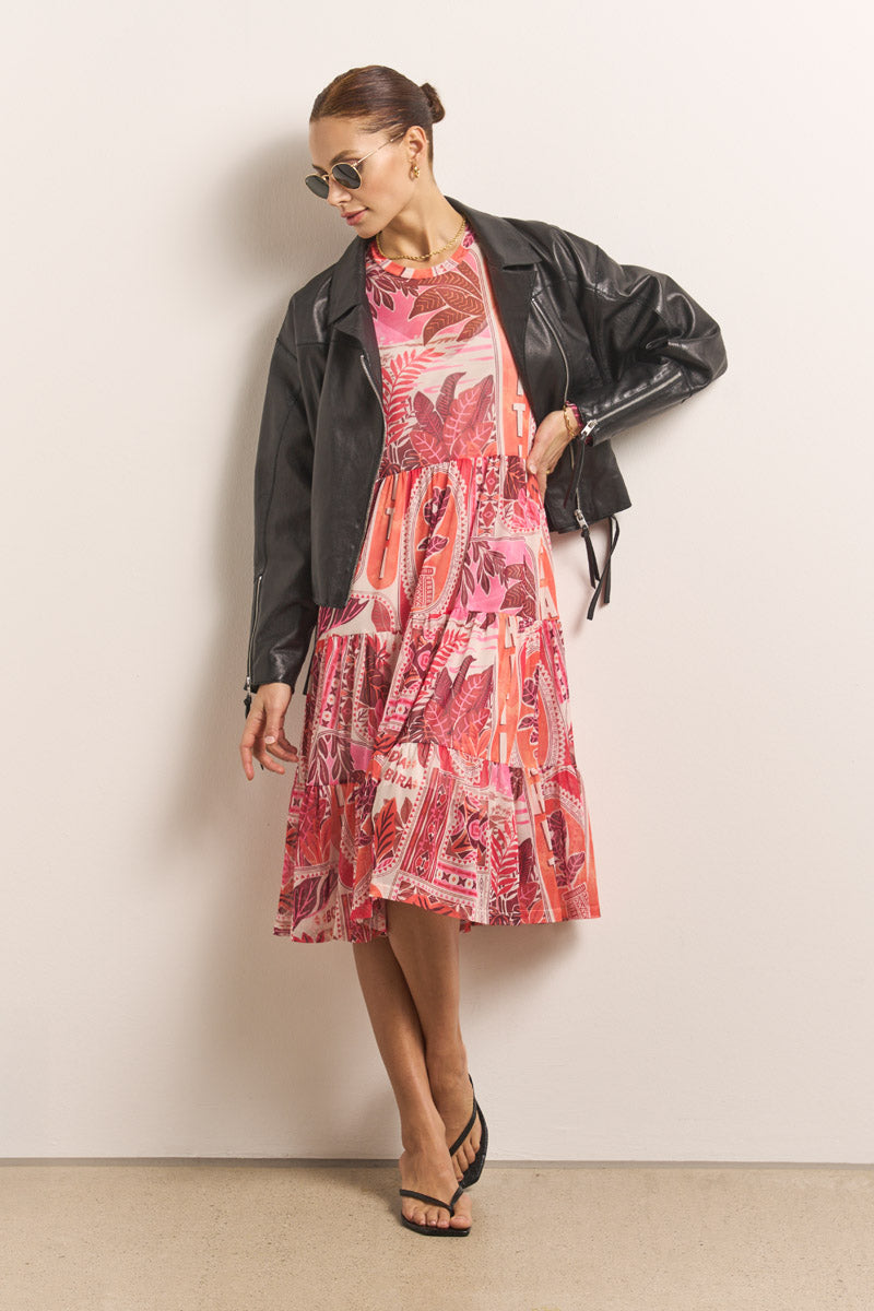 Eine Frau mit Princess Goes Hollywood-Sonnenbrille, schwarzer Lederjacke und knielangem rosa Kleid mit Tahiti-Print steht vor einer schlichten Wand, die Haare nach hinten gebunden, den Blick nach unten gerichtet, schwarze Sandalen tragend und einen Fuß vor den anderen gekreuzt.