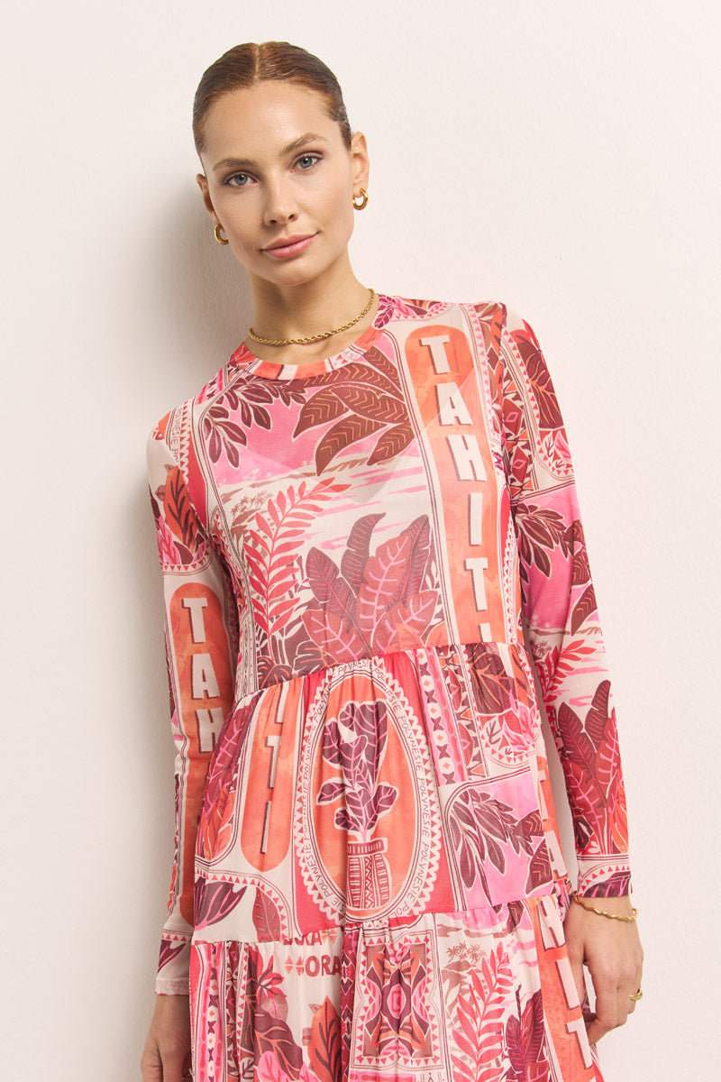 Eine Frau mit heller Haut und hochgestecktem Haar steht vor einer weißen Wand. Sie trägt ein langärmeliges Princess Goes Hollywood Tahiti-Print Mesh-Kleid mit tropischen Blättern und "TAHITI"-Aufdruck, dazu goldene Creolen und eine zarte goldene Halskette.