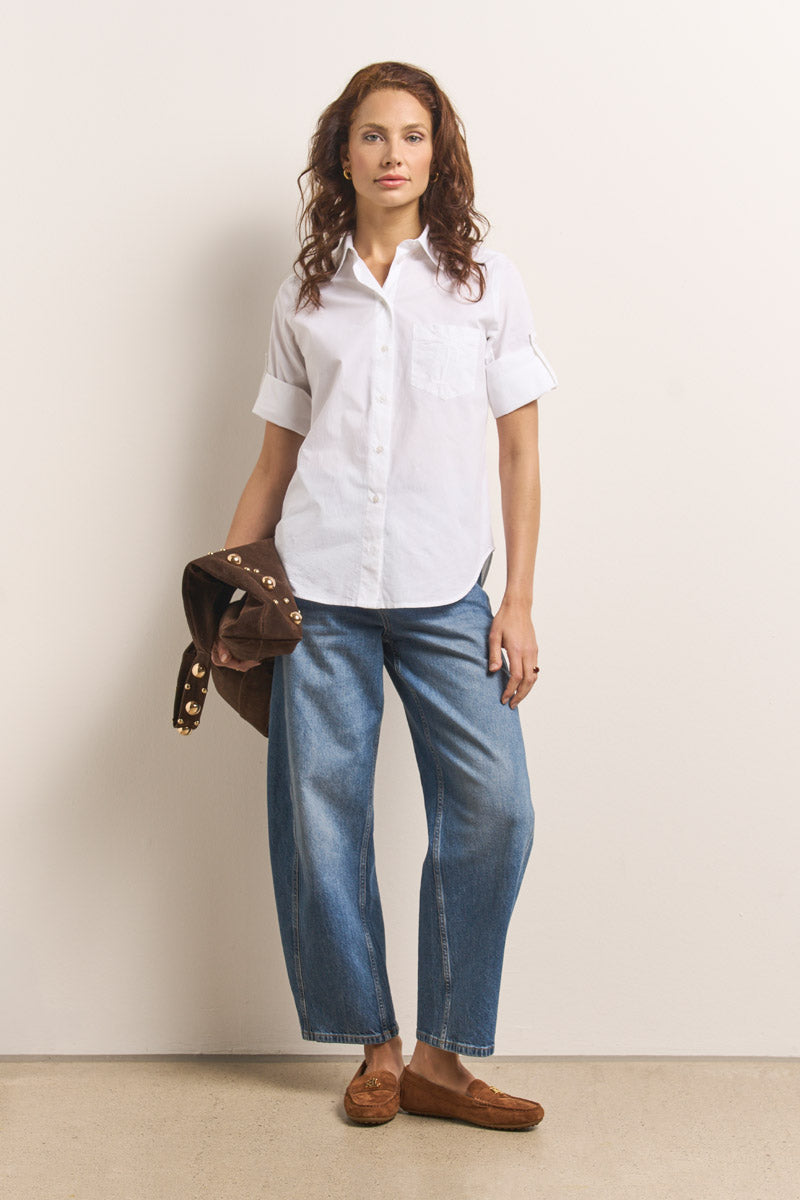 Eine Frau mit gewelltem braunem Haar steht an einer schlichten Wand und trägt eine weiße Hemdbluse aus Baumwollmix von Princess Goes Hollywood mit gekrempelten Ärmeln, weite Bluejeans und braune Slipper. Über dem linken Arm hält sie eine braune Jacke mit Goldknöpfen.