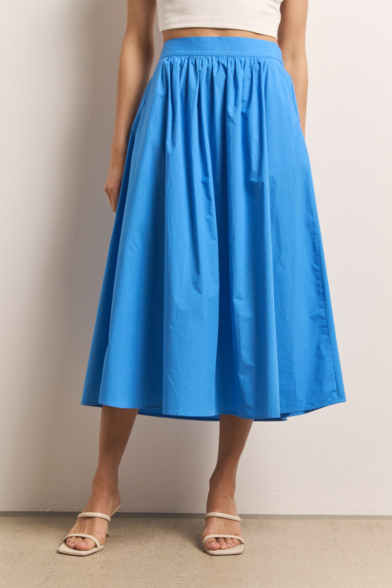 Eine Person trägt einen leuchtend blauen Baumwolle Midirock mit elastischem Bund von Princess Goes Hollywood, gestylt mit einem weißen Kurztop und beigen Absatzsandalen. Die Szene hat einen neutralen Hintergrund mit unifarbenen Wänden und Böden.