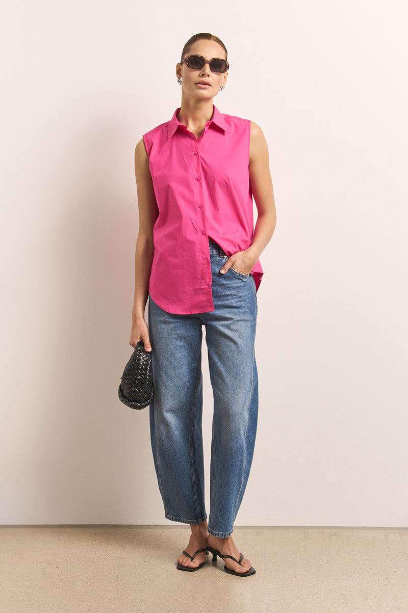 Eine Frau posiert vor einer schlichten Wand in einer leuchtend rosa ärmellosen Hemdbluse aus Baumwolle von Princess Goes Hollywood, weiten Bluejeans, schwarzen Flip-Flops, dunkler Sonnenbrille und einer kleinen schwarzen gewebten Handtasche. Ihr Haar ist zurückgebunden und eine Hand steckt in ihrer Tasche.