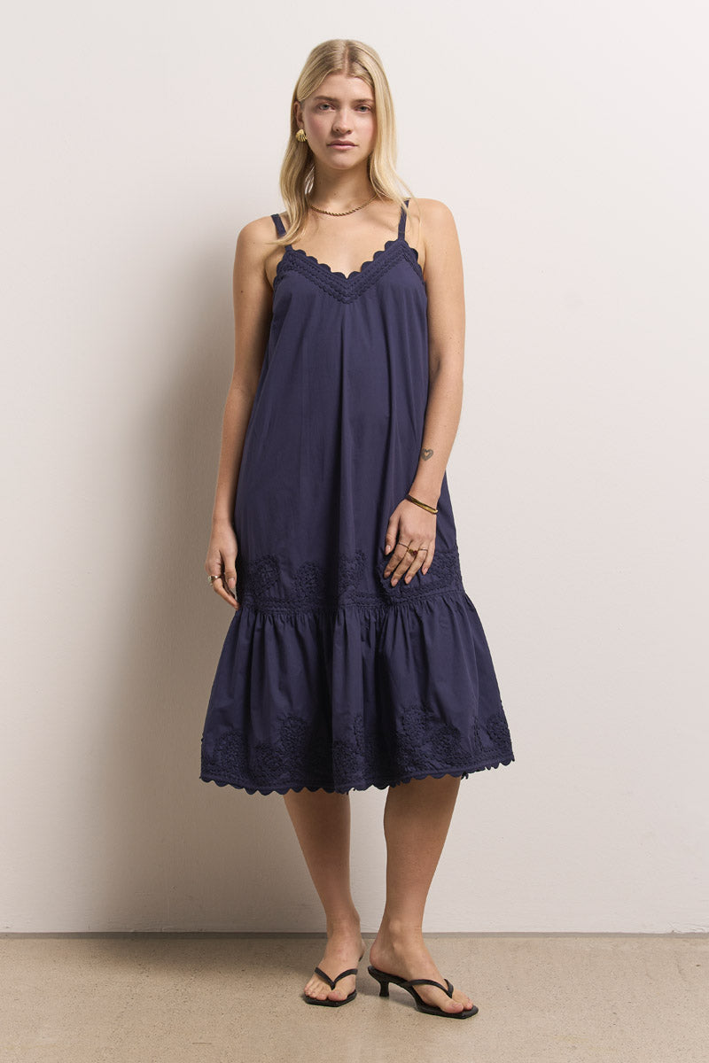 Eine Frau mit langen blonden Haaren steht vor einer schlichten Wand und trägt ein marineblaues Midikleid aus Baumwolle mit Spitzendetails und bestickten Wellenkanten von Princess Goes Hollywood. Dazu trägt sie eine mehrlagige goldene Halskette, ein Armband und schwarze Flip-Flops.