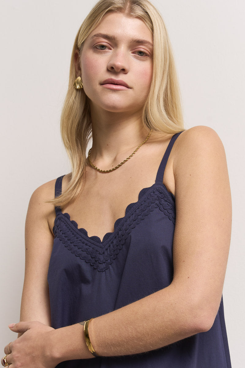 Eine junge Frau mit langen blonden Haaren trägt ein marineblaues Camisole mit Muschelmuster, goldene Creolen und eine goldene Halskette von Princess Goes Hollywood - ihr ruhiger, eleganter Look passt perfekt zu einem Princess Goes Hollywood Baumwolle Midikleid.