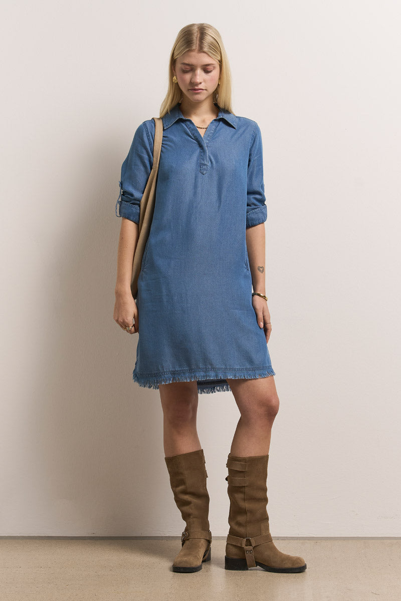 Eine Frau posiert vor einer schlichten Wand in einem blauen Princess Goes Hollywood Lyocell Blusenkleid mit Dreiviertelärmeln, Hemdkragen und ausgefranstem Saum, gepaart mit einer hellbraunen Umhängetasche, kniehohen Stiefeln aus braunem Wildleder und blondem Mittelscheitel.