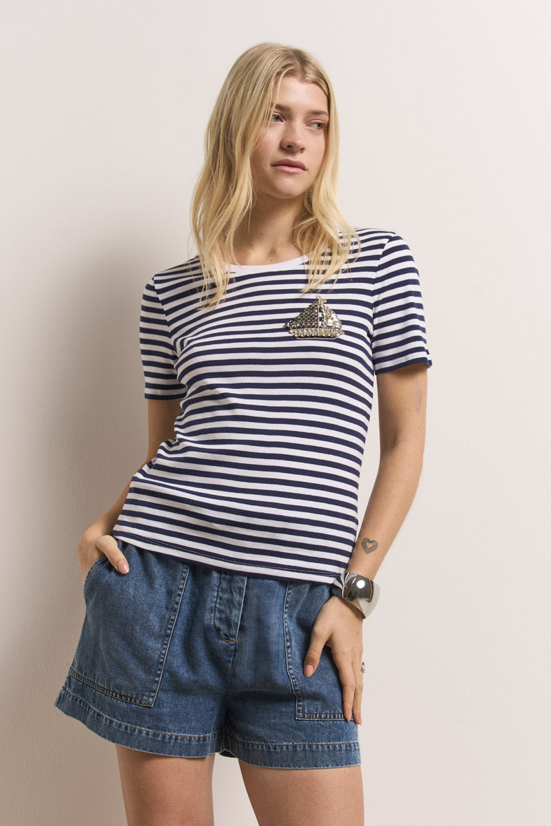 Eine Frau mit langen blonden Haaren, die ein marineblau-weiß gestreiftes kurzärmeliges Hemd und eine Jeansshorts aus Denim Paperbag-Bund von Princess Goes Hollywood trägt, steht an einer hellen Wand, hat die Hände in den Taschen und blickt nach links. Auf ihrem Arm ist eine Herz-Tätowierung zu sehen.