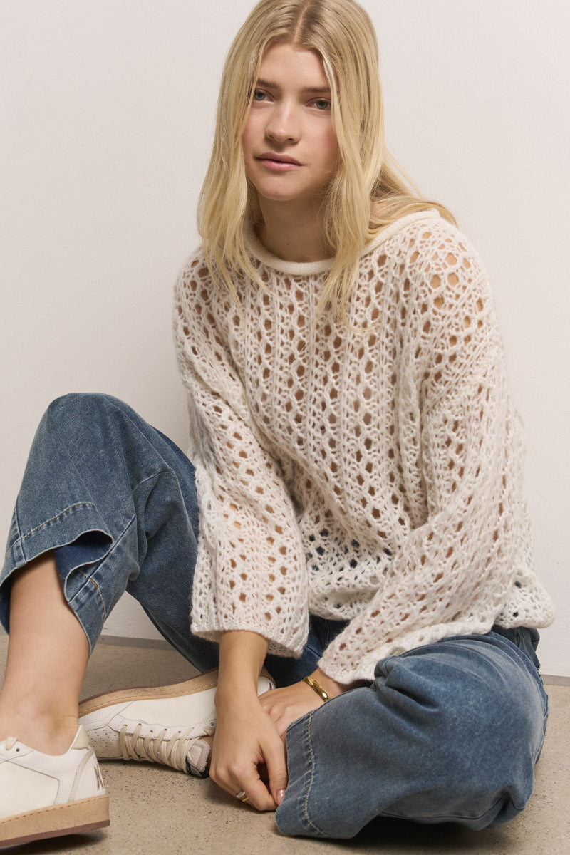 Eine junge Frau mit langen blonden Haaren sitzt an einer weißen Wand und trägt einen lockeren cremefarbenen offenen Strickpullover, eine blaue Paperbag-Bund Jeanshose aus Baumwolldenim von Princess Goes Hollywood und weiße Turnschuhe. Sie blickt sanft in die Kamera.