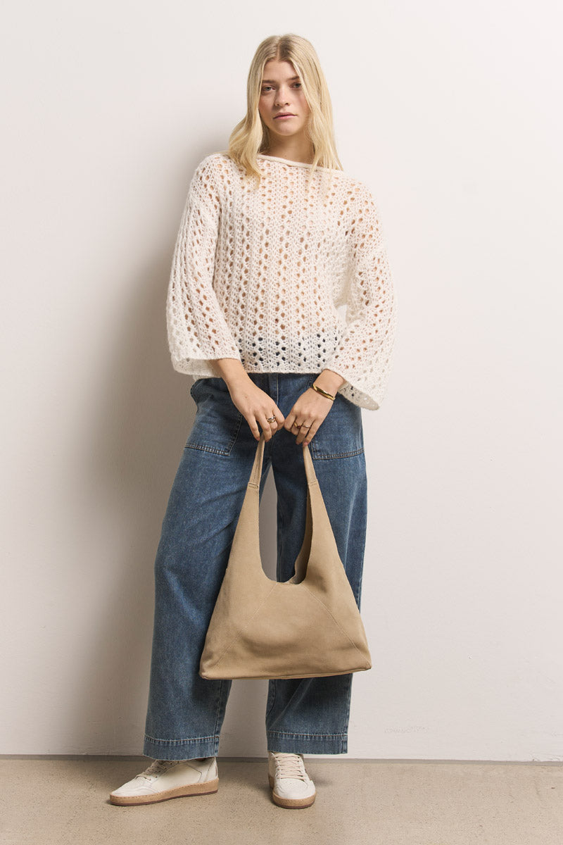 Eine Frau mit langen blonden Haaren steht an einer schlichten Wand und trägt einen lockeren weißen Häkelpulli von Princess Goes Hollywood über einem beigen Oberteil, eine Baumwolldenim-Jeanshose mit weitem Bein, weiße Turnschuhe und eine große beigefarbene Schlabbertasche.