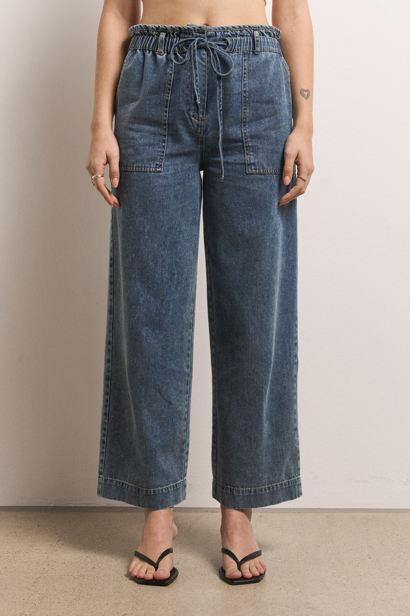 Eine Person steht vor einer hellen Wand in Princess Goes Hollywood in einer blauen Paperbag-Bund-Jeanshose mit hohem Bund, gerafftem Gummiband, Kordelzug vorne und großen Taschen. Schwarze Sandalen und ein kleines Herz-Tattoo auf dem linken Unterarm vervollständigen den Look.