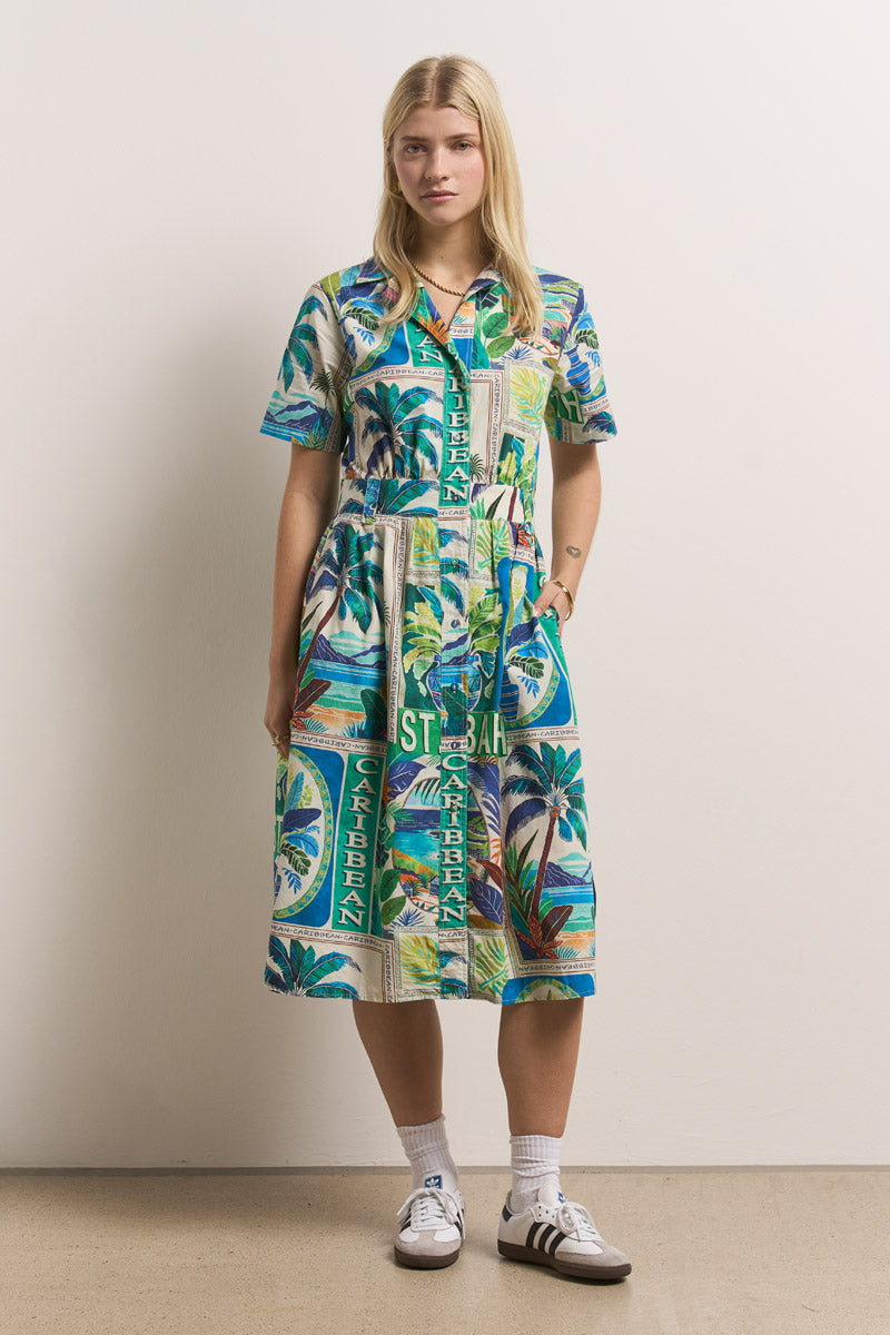 Eine Frau mit glattem blondem Haar trägt ein farbenfrohes Baumwoll-Midikleid mit St. Barth-Aufdruck von Princess Goes Hollywood mit Palmen und dem Schriftzug "ST BARTH CARIBBEAN", dazu weiße Socken und klassische weiße Adidas-Turnschuhe.