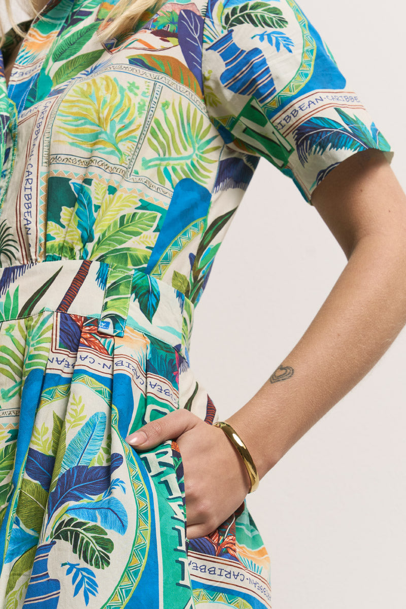Eine Frau, die vom Oberkörper bis zum Arm gezeigt wird, trägt ein farbenfrohes Princess Goes Hollywood Midikleid mit einem tropischen St. Barth-Aufdruck und "CARIBBEAN"-Aufklebern. Sie hat eine Hand in der Tasche, ein goldenes Armband und ein kleines Herz-Tattoo am Handgelenk.