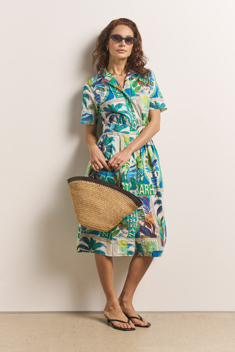Eine Frau mit gewellten braunen Haaren steht in einem farbenfrohen Princess Goes Hollywood St. Barth-Print-Midikleid aus Baumwolle vor einer schlichten Wand und strahlt mit braunen Sandalen, dunkler Sonnenbrille und einer geflochtenen Strohtasche einen entspannten Sommerstil aus.