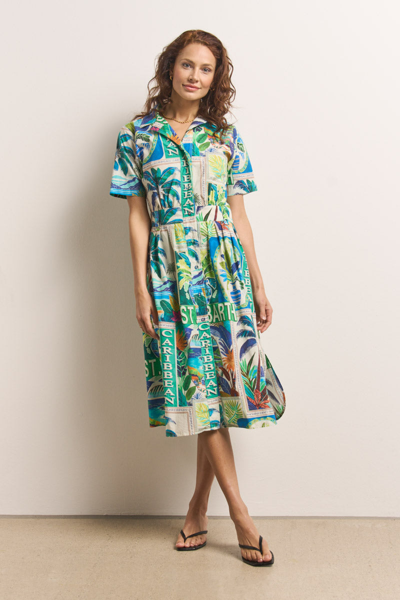 Eine Frau mit gewelltem kastanienbraunem Haar trägt ein farbenfrohes Midikleid aus Baumwolle mit St. Barth-Print von Princess Goes Hollywood, kurzen Ärmeln und tropischen Mustern. Sie lehnt an einer schlichten Wand, trägt Flip-Flops und lächelt sanft.