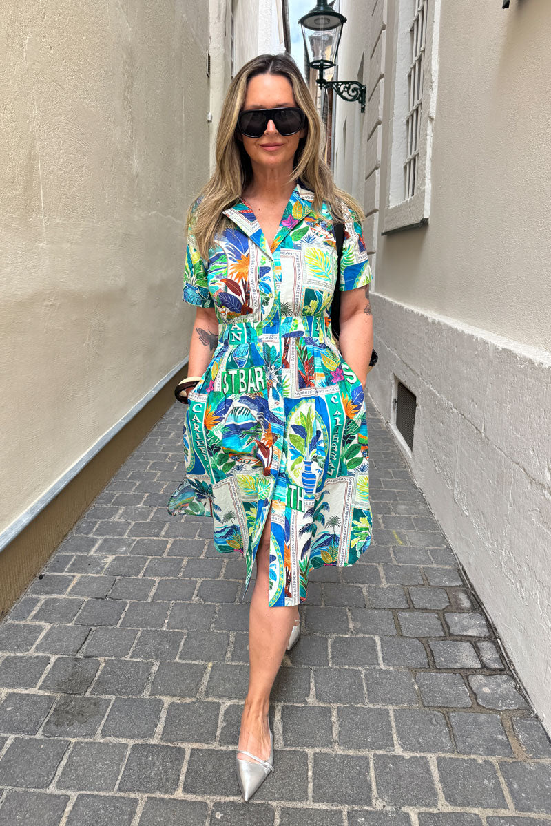 Eine Frau mit langen blonden Haaren und Sonnenbrille geht selbstbewusst in einem bunten Princess Goes Hollywood Baumwolle Midikleid mit St. Barth-Print und spitzen silbernen Absätzen eine kopfsteingepflasterte Gasse entlang, die Hände in den Taschen vor hellen Stadtmauern.