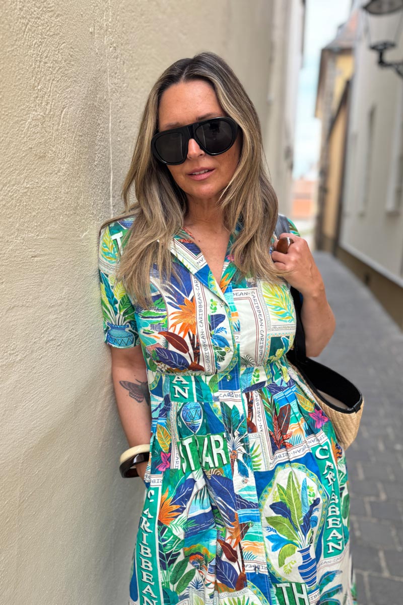 Eine Frau mit langen blonden Haaren trägt eine große schwarze Sonnenbrille und ein buntes Baumwoll-Midikleid mit karibischen Mustern im Princess Goes Hollywood St. Barth-Print. Sie steht vor einer sonnenbeschienenen beigen Wand, hält eine Strohtasche und trägt Armbänder.