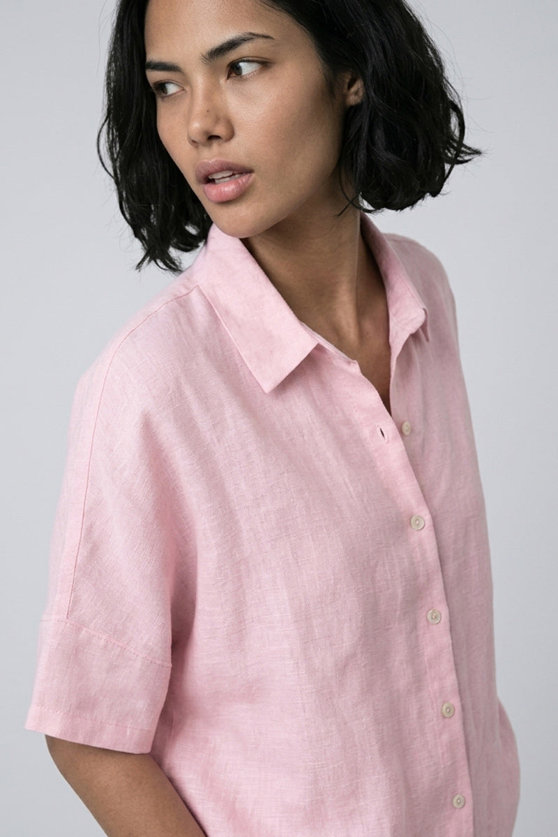 Eine Frau mit kurzen dunklen Haaren und heller Haut trägt eine hellrosa, kurzärmlige Oversize-Bluse aus Leinen von Princess Goes Hollywood. Sie blickt mit neutralem Blick nach links vor einem schlichten, hellgrauen Hintergrund.