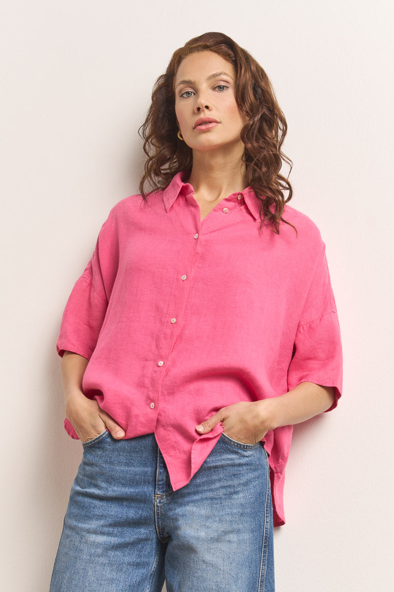 Eine Frau mit gewelltem braunem Haar trägt eine leuchtend rosa Oversize-Bluse aus Leinen mit Hemdkragen von Princess Goes Hollywood und blaue Jeans. Sie steht vor schlichtem Hintergrund, Hände in den Taschen, und blickt selbstbewusst in die Kamera.