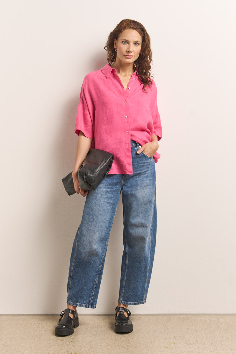 Eine Frau steht vor einer weißen Wand und trägt eine übergroße rosa Bluse mit Hemdkragen von Princess Goes Hollywood, gepaart mit weit geschnittenen Bluejeans und schwarzen Plateausandalen. Sie hält eine große schwarze Clutch in der Hand und blickt mit einem leichten Lächeln in die Kamera.