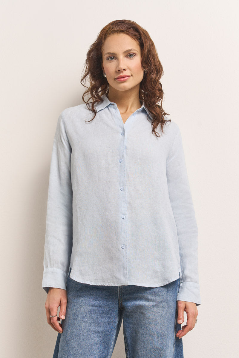 Eine Frau mit gewelltem, schulterlangem kastanienbraunem Haar trägt eine hellblaue Princess Goes Hollywood Damen Leinen Hemdbluse und blaue Jeans. Sie steht mit dem Gesicht nach vorne vor einem cremefarbenen Hintergrund und hat die Arme entspannt an den Seiten verschränkt.