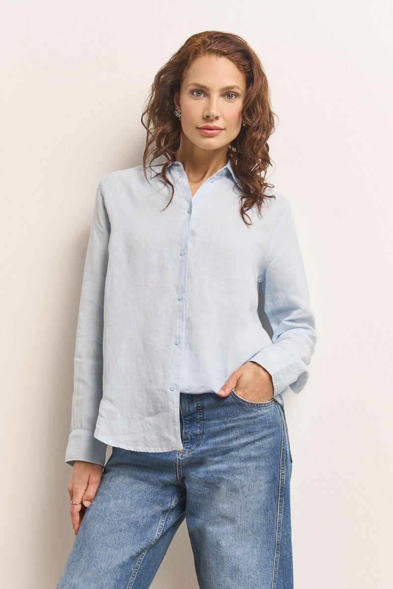 Eine Frau mit gewelltem kastanienbraunem Haar steht vor einer weißen Wand und trägt eine hellblaue Princess Goes Hollywood Damen Bluse mit Langarm und Blue Jeans. Eine Hand steckt in ihrer Tasche, während sie mit entspanntem Blick direkt in die Kamera schaut.