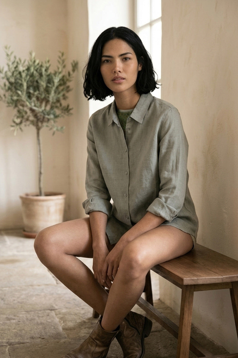 Eine Frau mit kurzen, dunklen Haaren sitzt auf einer Holzbank in einem Innenraum. Sie trägt eine Princess Goes Hollywood Leinenhemdbluse in Hellgrau und braune Stiefeletten. Hinter ihr steht eine Topfpflanze vor einer cremefarbenen Wand, die von natürlichem Fensterlicht sanft beleuchtet wird.