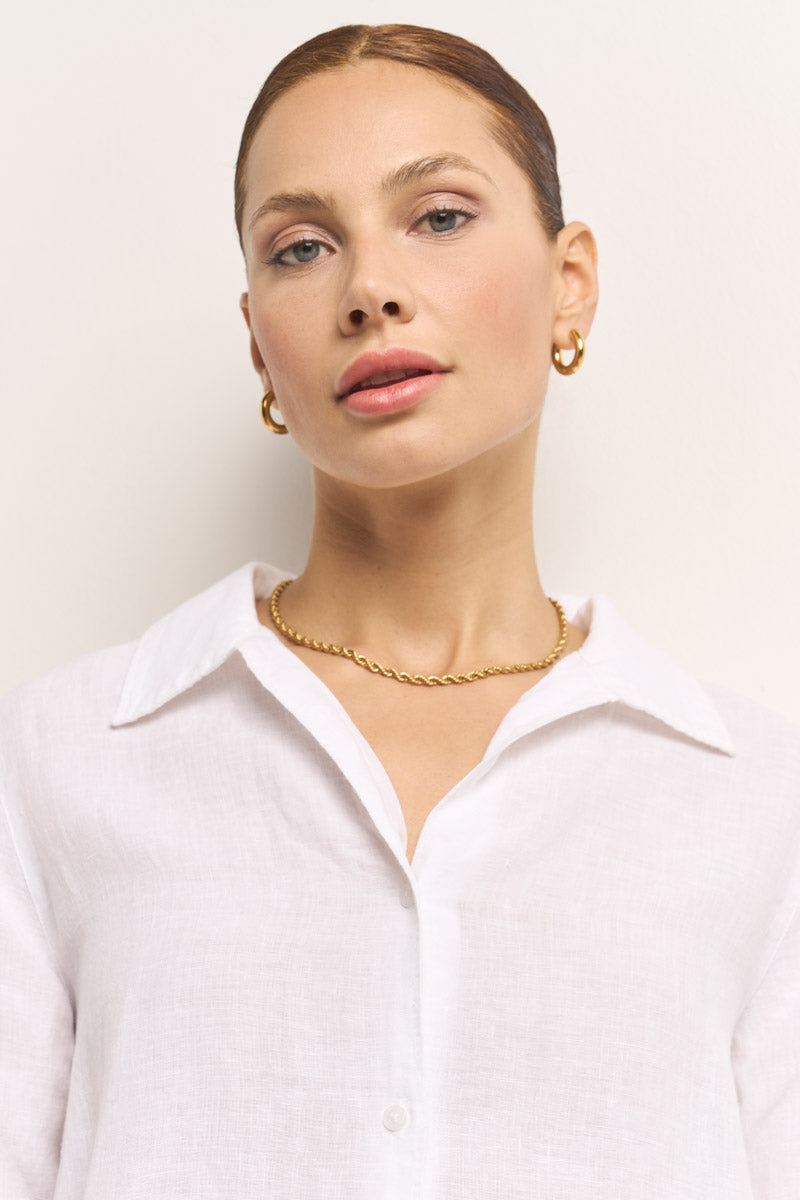 Eine Frau mit heller Haut und zurückgekämmten braunen Haaren blickt in die Kamera. Sie trägt Goldschmuck und ein weißes Leinenhemd mit einem Kragen aus Princess Goes Hollywood. Der Hintergrund ist einfarbig und hell.