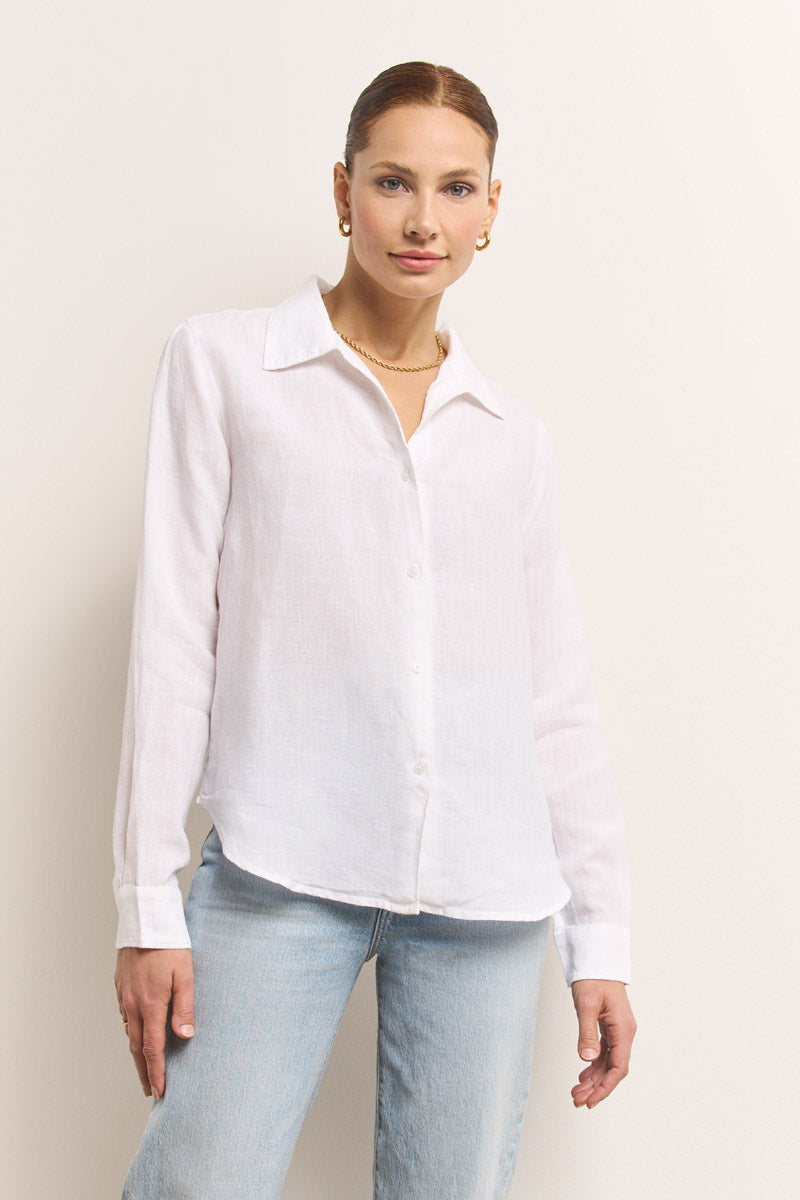 Eine Frau mit hellbraunem, nach hinten gestecktem Haar trägt ein weißes Leinenhemd mit Hemdkragen und langen Ärmeln von Princess Goes Hollywood, gepaart mit hellblauen Jeans. Sie steht vor einer cremefarbenen Wand und trägt goldene Reifen und eine goldene Halskette.