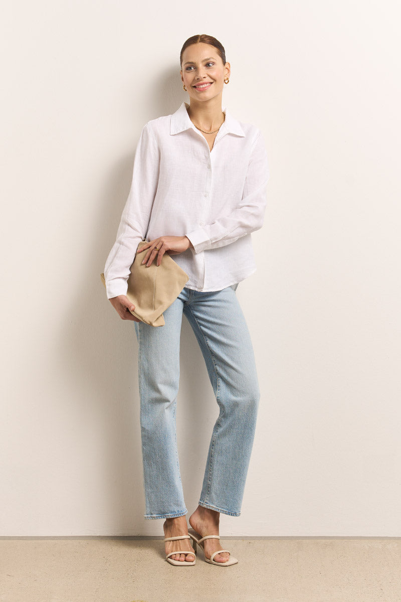 Eine Frau lächelt gegen eine helle Wand. Sie trägt eine Princess Goes Hollywood weiße Leinen-Hemdbluse mit langen Ärmeln, hellblaue Jeans mit geradem Bein, weiße Sandaletten mit Absatz, eine beigefarbene Clutch und goldene Ohrringe und Halskette als Accessoires.