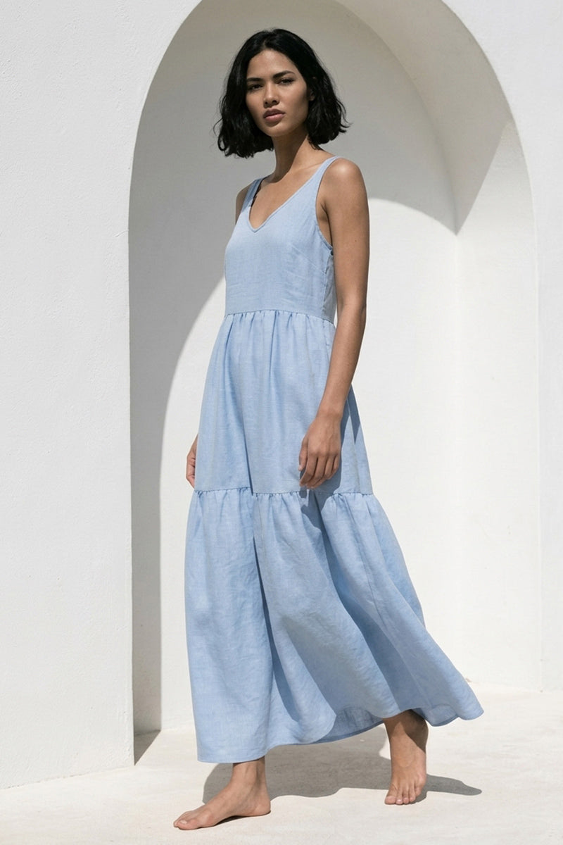 Eine Frau mit kurzen dunklen Haaren steht barfuß vor einer weißen gewölbten Wand und trägt ein hellblaues Princess Goes Hollywood Midikleid aus Leinen mit Seitentaschen. Das ärmellose Kleid mit V-Ausschnitt hat einen gestuften Rock, der in einer hellen, minimalen Szenerie mit weichem natürlichem Licht gezeigt wird.
