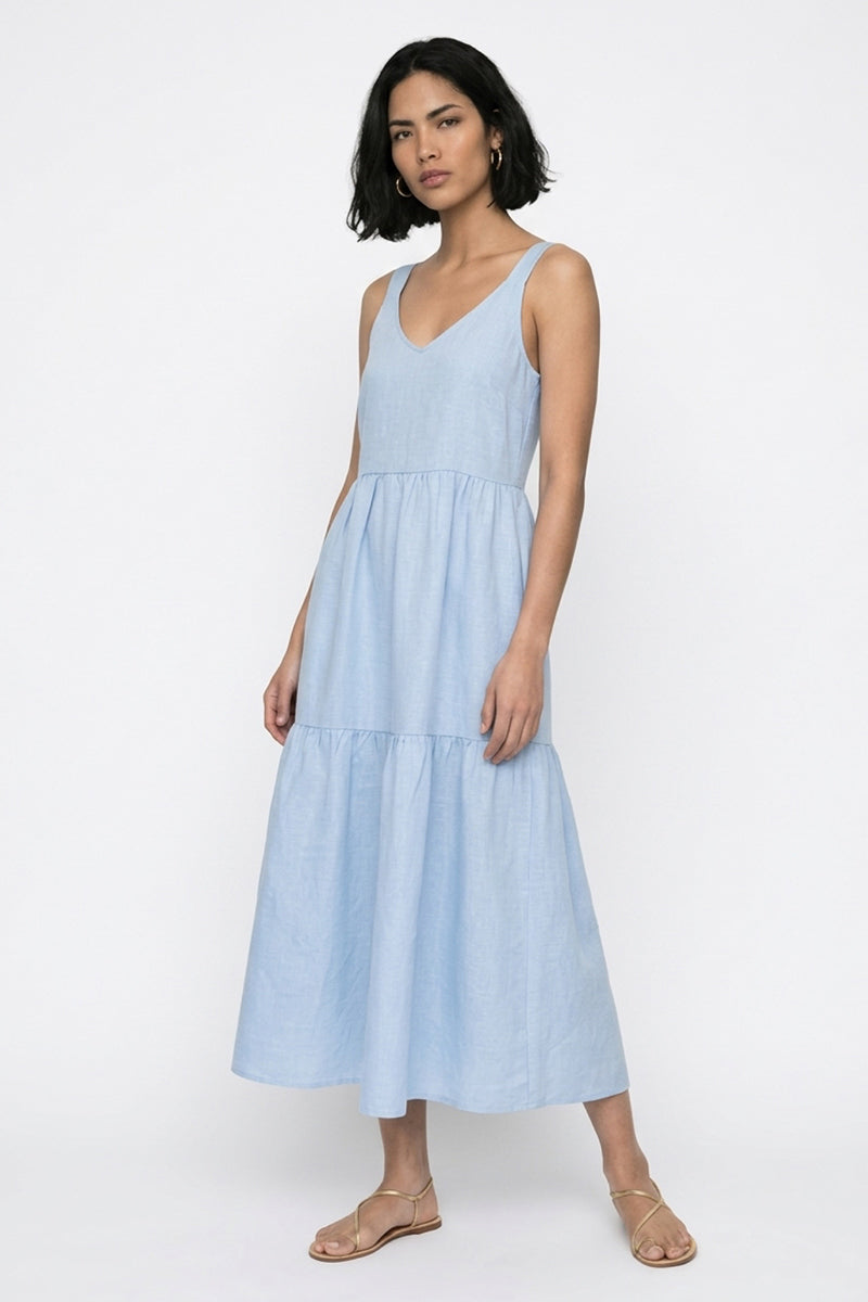 Eine Frau mit schulterlangem dunklem Haar trägt ein hellblaues Princess Goes Hollywood Kleid mit Trägern mit V-Ausschnitt und gestuftem, ausgestelltem Kleidrock. Sie steht in flachen Riemchensandalen vor einem weißen Hintergrund, die Arme entspannt, der Gesichtsausdruck neutral.