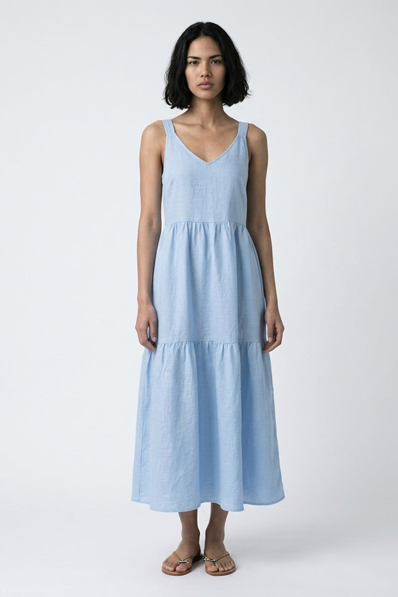 Eine Frau mit einem Bob-Haarschnitt trägt ein hellblaues Princess Goes Hollywood Midikleid Leinen mit V-Ausschnitt und Stufenrock, gepaart mit flachen Sandalen. Sie steht mit dem Gesicht nach vorne vor einem schlichten hellgrauen Hintergrund, ihr Gesichtsausdruck ist neutral.