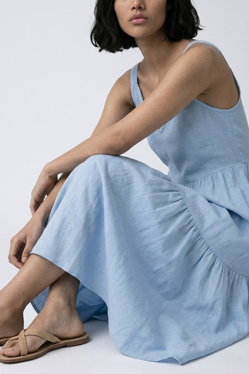Eine Frau mit kurzem, dunklem Haar und heller Haut trägt ein hellblaues Midikleid aus Leinen mit Trägern von Princess Goes Hollywood. Sie sitzt mit einem angehobenen Knie und kombiniert das ausgestellte Kleid mit flachen gewebten beigen Sandalen vor einem schlichten grauen Hintergrund.