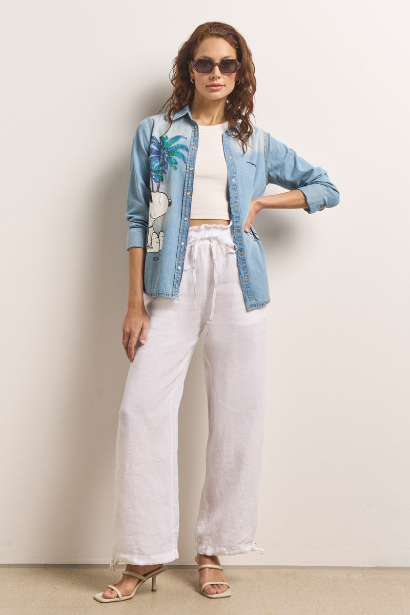 Eine Frau posiert selbstbewusst mit einer Princess Goes Hollywood-Sonnenbrille, einem hellblauen Jeanshemd mit Snoopy- und Palmengrafik, einem weißen Crop-Top, einer weiten Leinenhose mit elastischem Bund und Kordelzug und beigen hochhackigen Sandalen. Eine Hand in der Tasche.