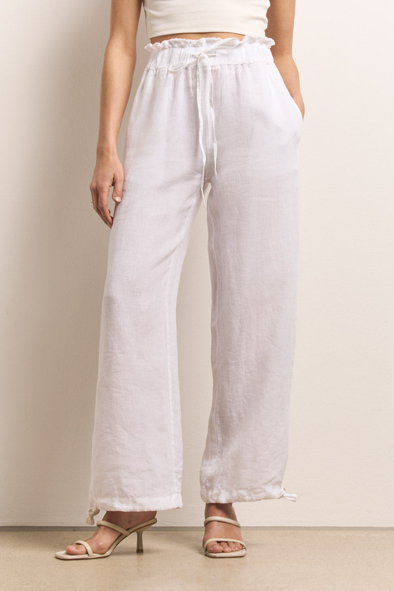 Die Person trägt eine weiße Leinenhose mit elastischem Bund und Kordelzug von Princess Goes Hollywood, dazu ein weißes, kurzes Oberteil und Sandalen mit leichten Absätzen und steht mit einer Hand in der Tasche vor einer neutralen Wand.