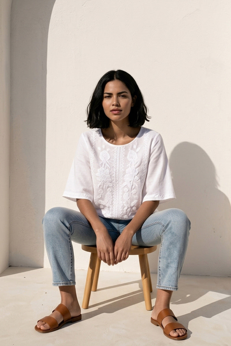Eine Frau mit Bobfrisur sitzt auf einem Holzhocker vor einer weißen Wand. Sie trägt eine Princess Goes Hollywood weiße Leinenbluse mit zarter Stickerei, hellblaue Jeans und braune Gleitsandalen. Das Sonnenlicht wirft weiche Schatten, während sie ruhig nach vorne schaut.