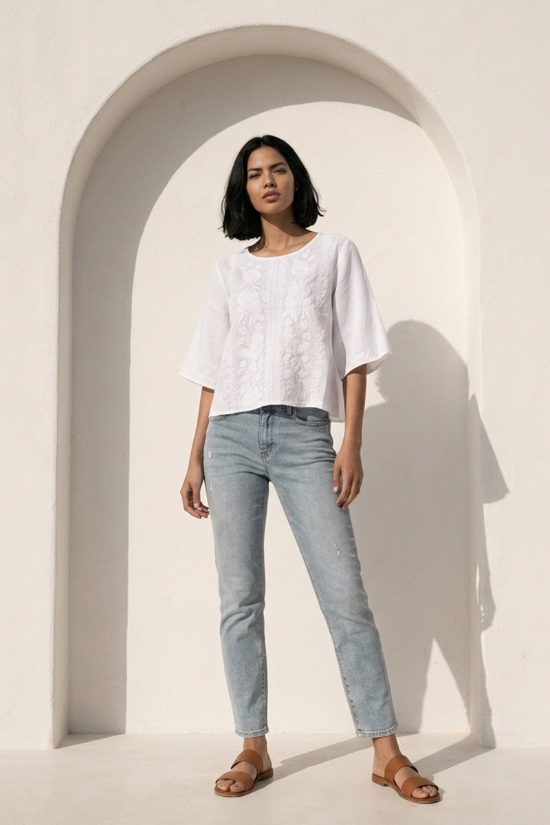 Eine Frau mit mittellangem Haarschnitt steht in einer lockeren Princess Goes Hollywood Leinen Bluse mit Stickerei, hellblauen Jeans und braunen Sandalen vor einer weißen gewölbten Wand. Das Sonnenlicht wirft ihren Schatten nach rechts, was den minimalistischen, modernen Eindruck noch verstärkt.