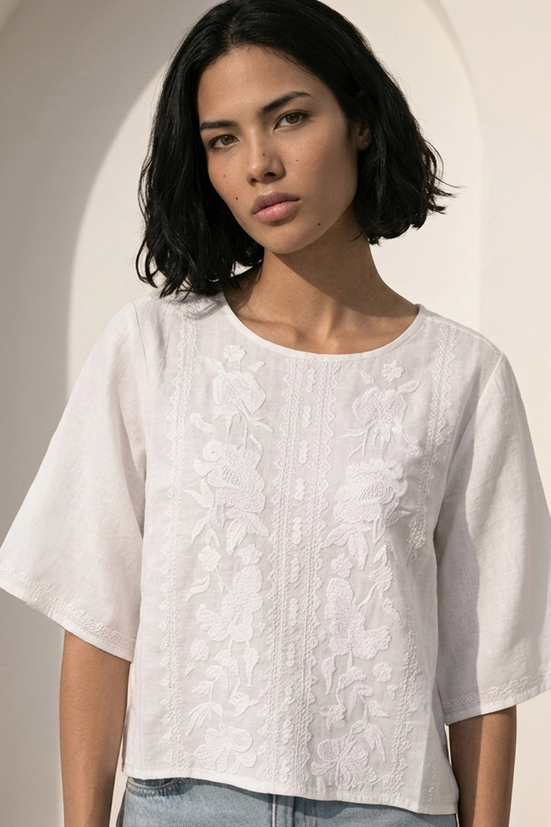 Eine Frau mit kinnlangem schwarzem Haar trägt eine Princess Goes Hollywood lockere Leinenbluse mit Rundhalsausschnitt und filigraner Stickerei, steht ruhig im sanften Sonnenlicht vor einem hellen gewölbten Hintergrund und blickt in die Kamera.