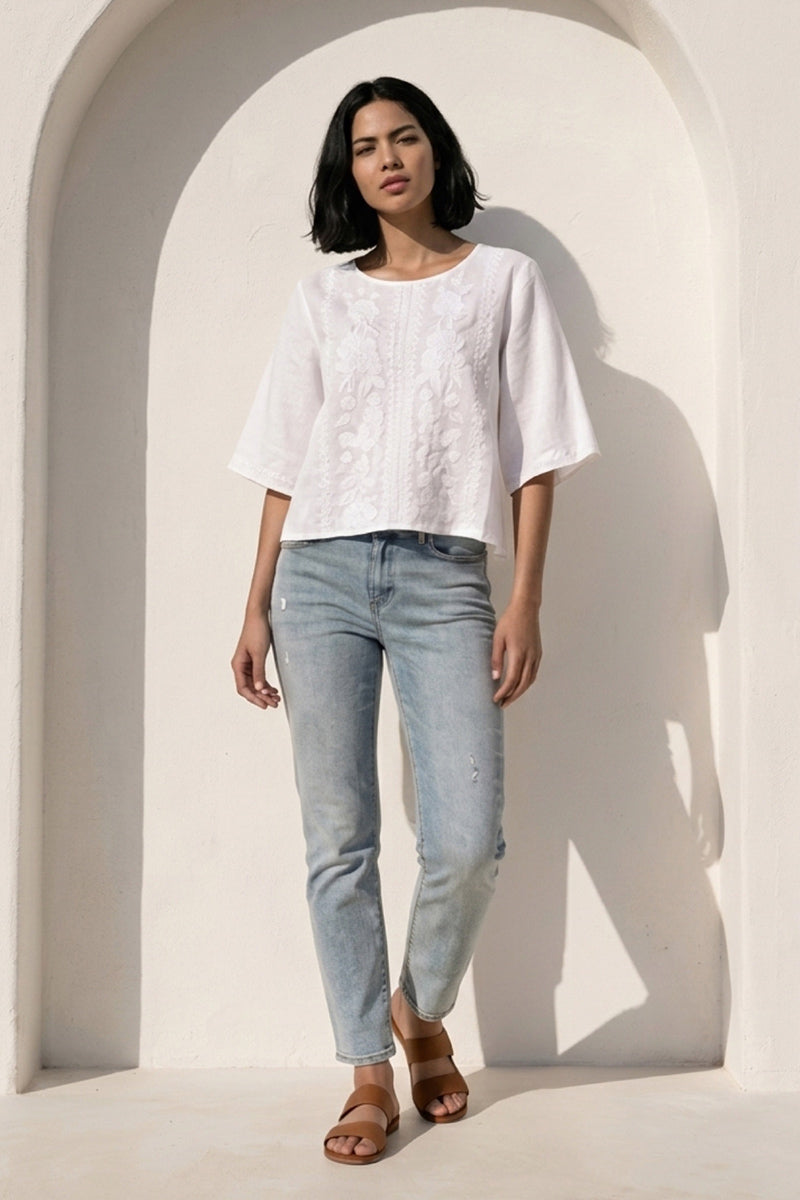 Eine Frau mit schulterlangem, dunklem Haar steht an einer weißen, gewölbten Wand. Sie trägt eine bestickte Leinenbluse von Princess Goes Hollywood, hellblaue Jeans und braune Sandalen. Sanftes Sonnenlicht wirft weiche Schatten, während sie mit neutraler Miene nach vorne blickt.