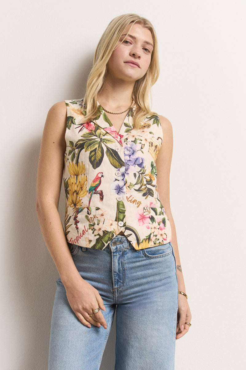 Eine Frau mit langen blonden Haaren trägt ein ärmelloses, mit bunten Blumen und Vögeln bedrucktes Button-up-Top mit Tropical-Print von Princess Goes Hollywood, gepaart mit hellblauen, hoch taillierten Jeans. Sie steht selbstbewusst vor einer schlichten weißen Wand, die linke Hand in der Tasche.