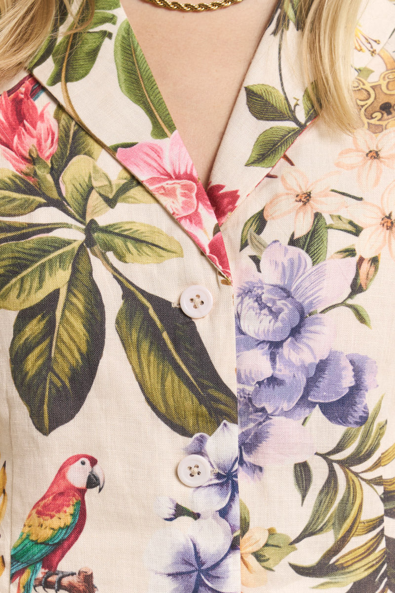 Eine Nahaufnahme eines Princess Goes Hollywood-Hemdes mit Tropical-Print mit großen grünen Blättern, roten und lila Blumen und einem roten Papagei. Das Hemd hat weiße Knöpfe und ist mit blonden Haaren, einer goldenen Halskette und einer Leinen Weste gestylt.