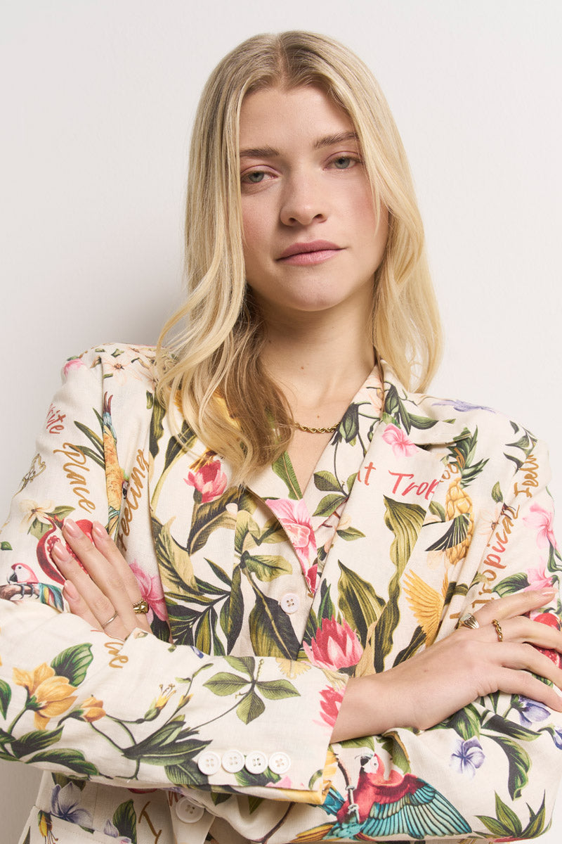 Eine Frau mit langen blonden Haaren und neutralem Gesichtsausdruck steht mit verschränkten Armen vor einem weißen Hintergrund und trägt einen farbenfrohen geblümten Damen Leinen Blazer von Princess Goes Hollywood und hellrosa Lippenstift.