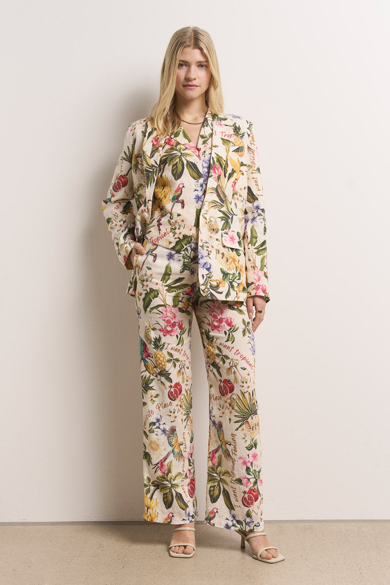 Eine Frau steht vor einer schlichten Wand in einem cremefarbenen Princess Goes Hollywood Leinenblazer und einer farbenfrohen Hose mit weitem Bein und tropischen Motiven, die bunte Pflanzen und Vögel zeigen - gestylt mit Sandalen und einem entspannten, selbstbewussten Look.