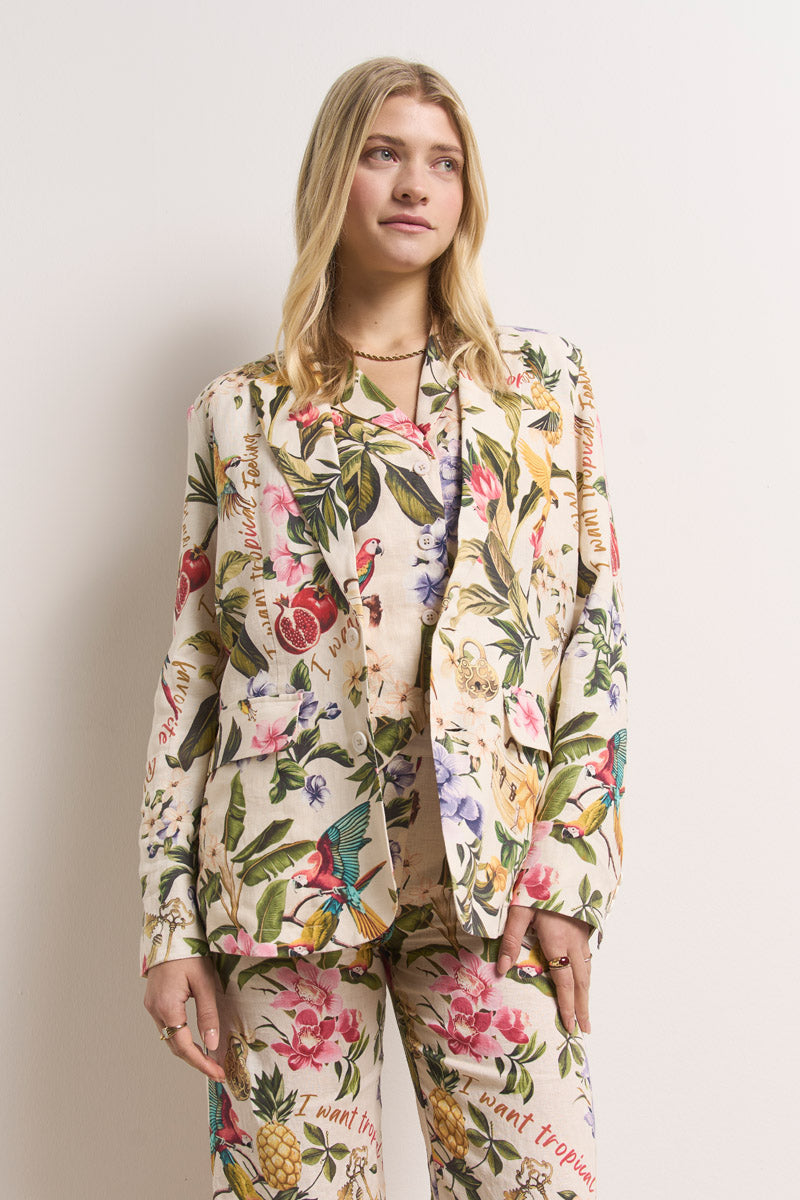 Eine Frau mit langen blonden Haaren steht vor einer weißen Wand und trägt einen Leinenblazer mit tropischem Muster und eine passende Hose von Princess Goes Hollywood. Ihr cremefarbener Anzug für Damen zeigt lebhafte Vögel, Früchte und bunte Blumen.