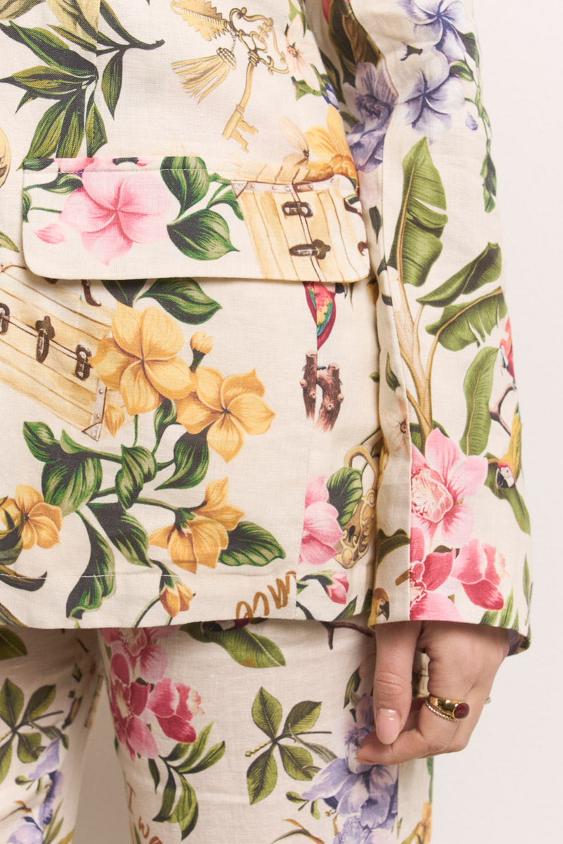 Nahaufnahme einer Person, die einen cremefarbenen Leinenblazer von Princess Goes Hollywood mit einem leuchtenden tropischen Druck in Gelb, Rosa, Lila und Grün, einer Klappentasche, goldenen Schlüssel- und Gepäckakzenten und zwei goldenen Ringen an der Hand trägt.