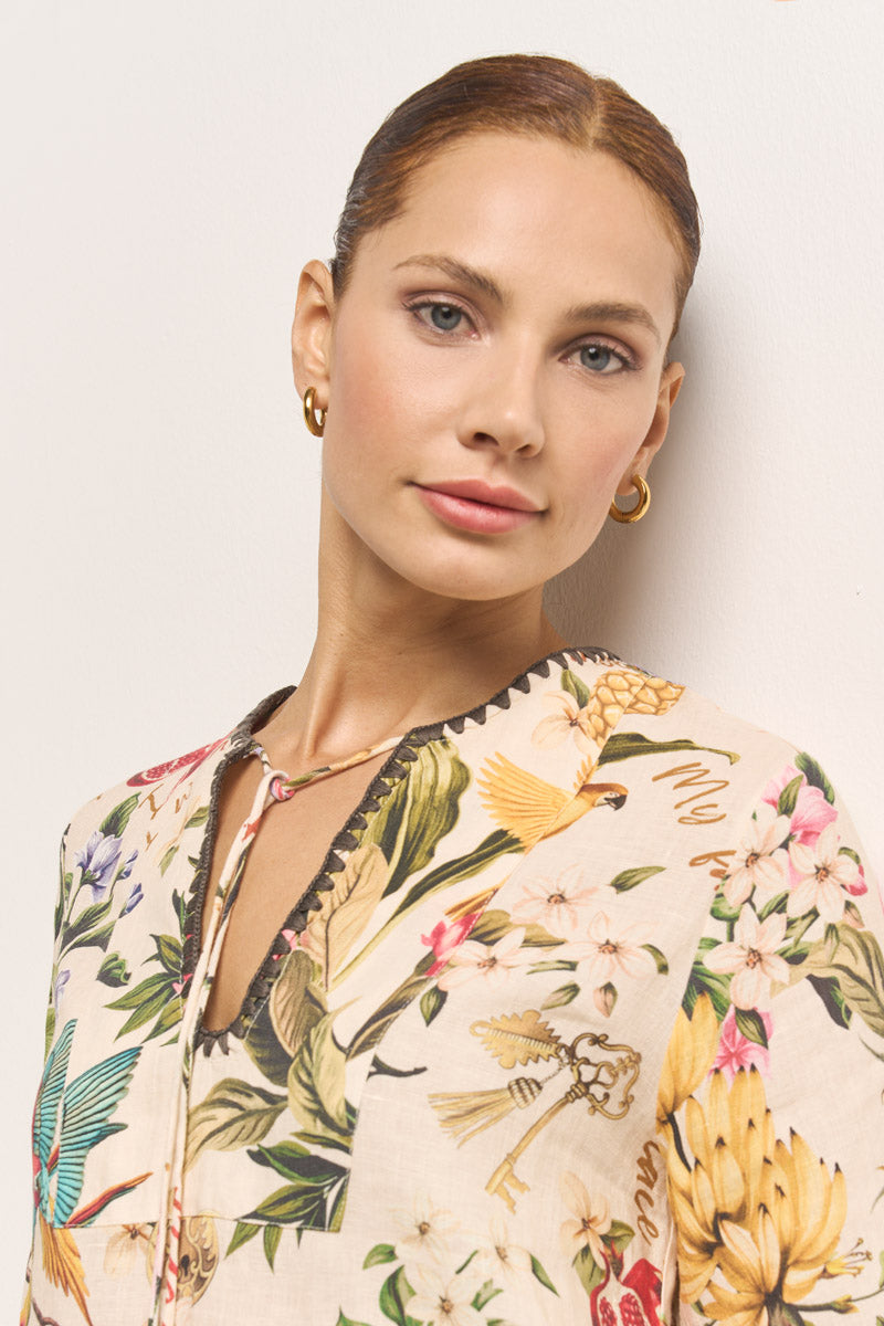 Eine hellhäutige Frau mit blauen Augen steht vor einer weißen Wand und trägt eine helle Leinen-Tunika mit tropischem Aufdruck und V-Ausschnitt von Princess Goes Hollywood. Ihr braunes Haar ist zurückgesteckt und sie trägt goldene Ohrringe.