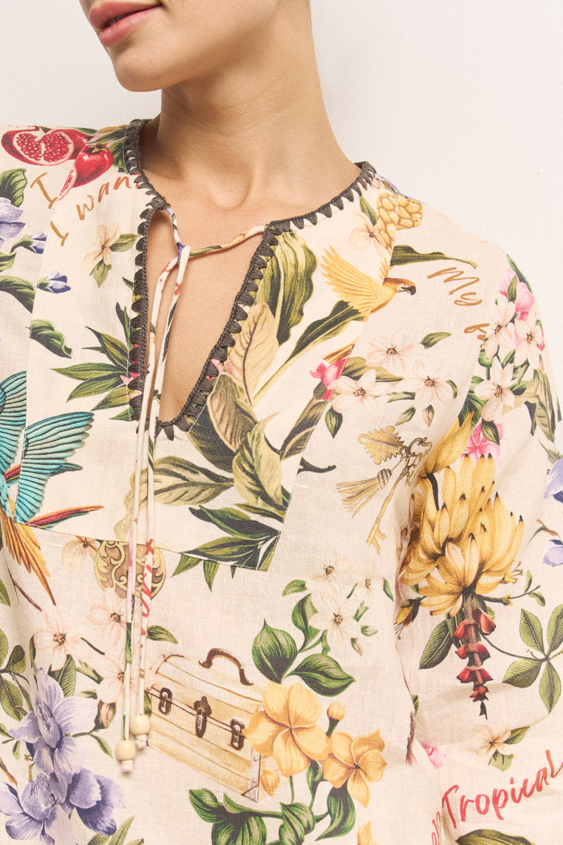 Eine Frau trägt eine Princess Goes Hollywood florale Tropical-Print Leinen Tunika mit Bananen, Vögeln und Blumen. Die Bluse hat einen V-Ausschnitt mit Schlitz und perlengeschmücktem Bindeband. Nur ihr unteres Gesicht und ihr oberer Oberkörper sind vor einem unifarbenen Hintergrund zu sehen.
