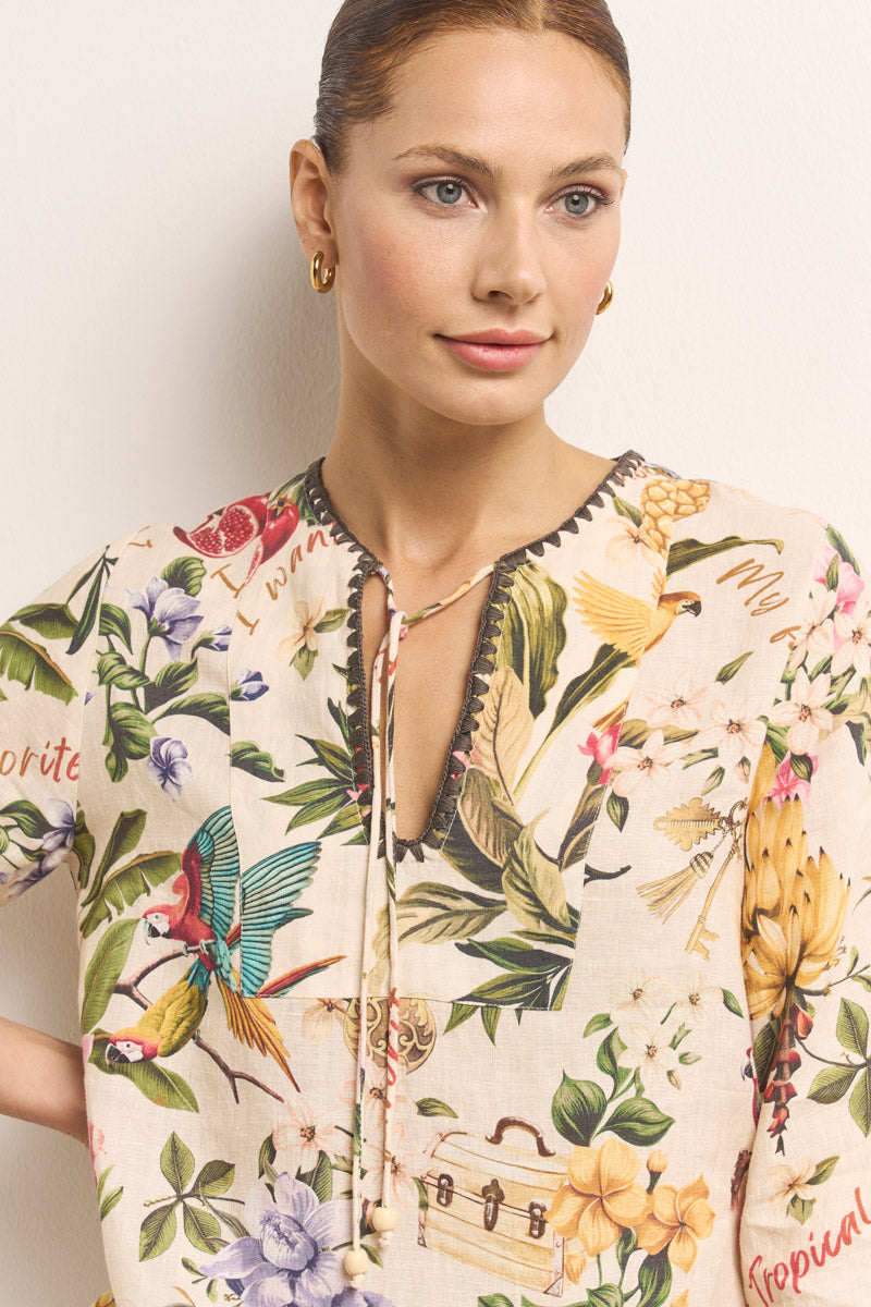 Eine hellhäutige, blauäugige Frau trägt eine tropisch bedruckte Princess Goes Hollywood Leinen-Tunika mit V-Ausschnitt, auf der Papageien, Blumen und Früchte abgebildet sind. Ihr braunes Haar ist zurückgesteckt, goldene Reifen glänzen, und sie trägt eine rosa Lippe auf neutralem Hintergrund.