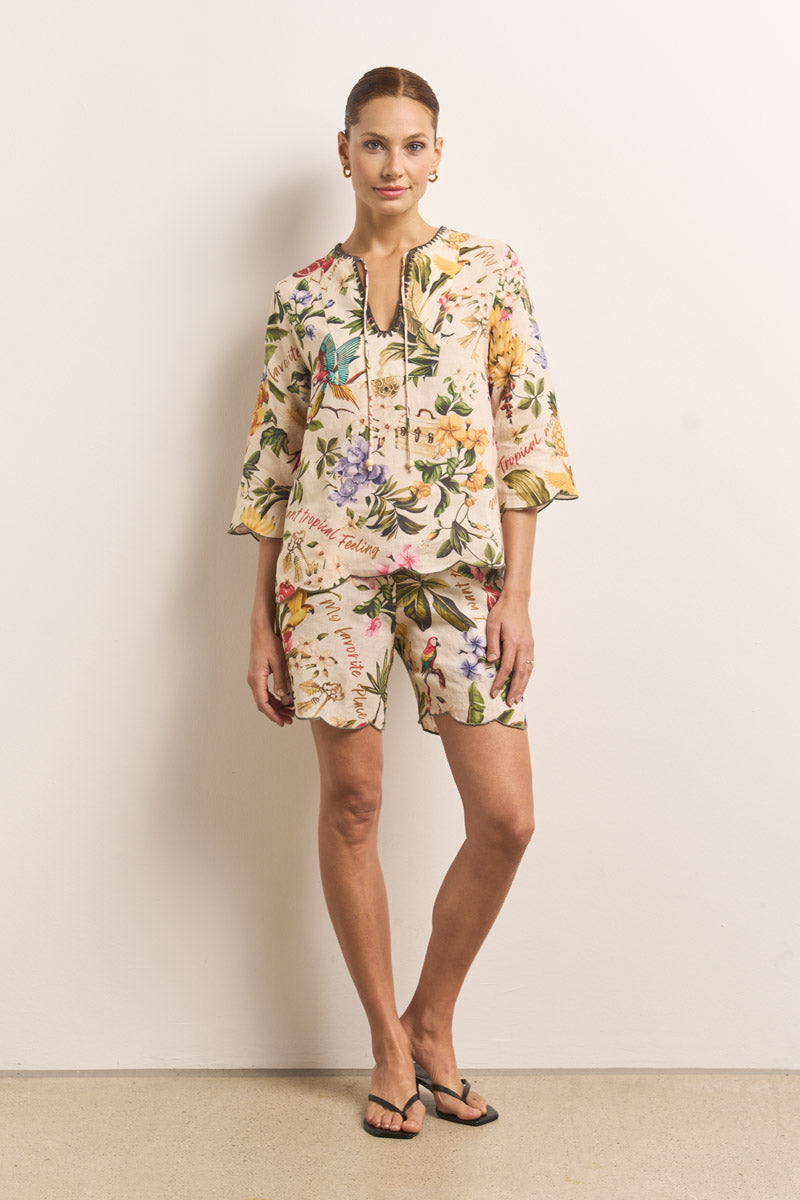 Eine Frau posiert lässig vor einer schlichten Wand in einem passenden hellen Oberteil mit tropischem Aufdruck und Shorts von Princess Goes Hollywood, mit zurückgebundenem Haar, Ohrringen, schwarzen Flip-Flops und einer Hand in der Tasche.