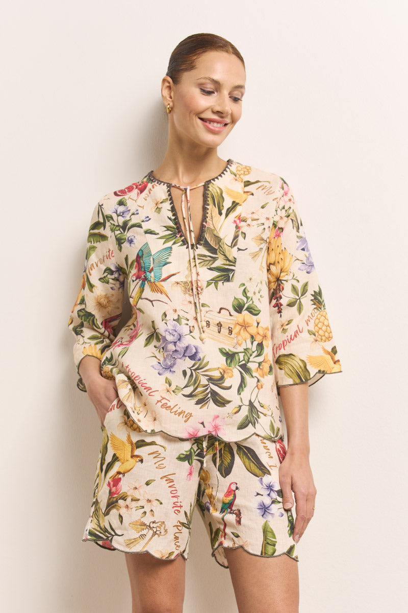 Eine Frau mit hochgestecktem Haar lächelt in einem beigefarbenen T-Shirt mit tropischem Druck und Shorts mit bunten Blumen und Vögeln von Princess Goes Hollywood. Das Shirt im Leinen-Tunika-Stil hat einen V-Ausschnitt und Dreiviertel-Ärmel. Sie steht vor einer cremefarbenen Wand.