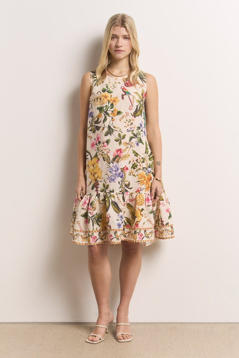 Eine Frau mit glattem, hellem Haar und neutralem Gesichtsausdruck steht vor einer schlichten, cremefarbenen Wand. Sie trägt ein ärmelloses Kurzkleid mit tropischem Aufdruck von Princess Goes Hollywood, dazu helle Sandalen und die Arme locker an der Seite.