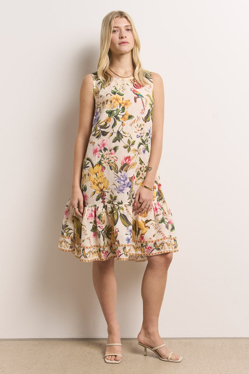 Eine Frau mit hellem Haar trägt ein ärmelloses, knielanges Sommerkleid mit tropischem Aufdruck von Princess Goes Hollywood. Das Kleid hat einen gerüschten Saum und sie trägt dazu Sandalen mit offenen Absätzen. Sie steht vor einer schlichten hellen Wand und schaut leicht zur Seite.